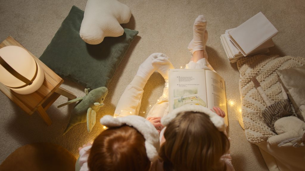 Two children in pajamas reading a book together at bedtime with soft lighting and toys around.